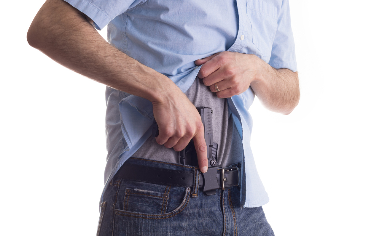 Person demonstrating appendix carry with a handgun in an inside-the-waistband holster positioned at the front of the waistband under a shirt.