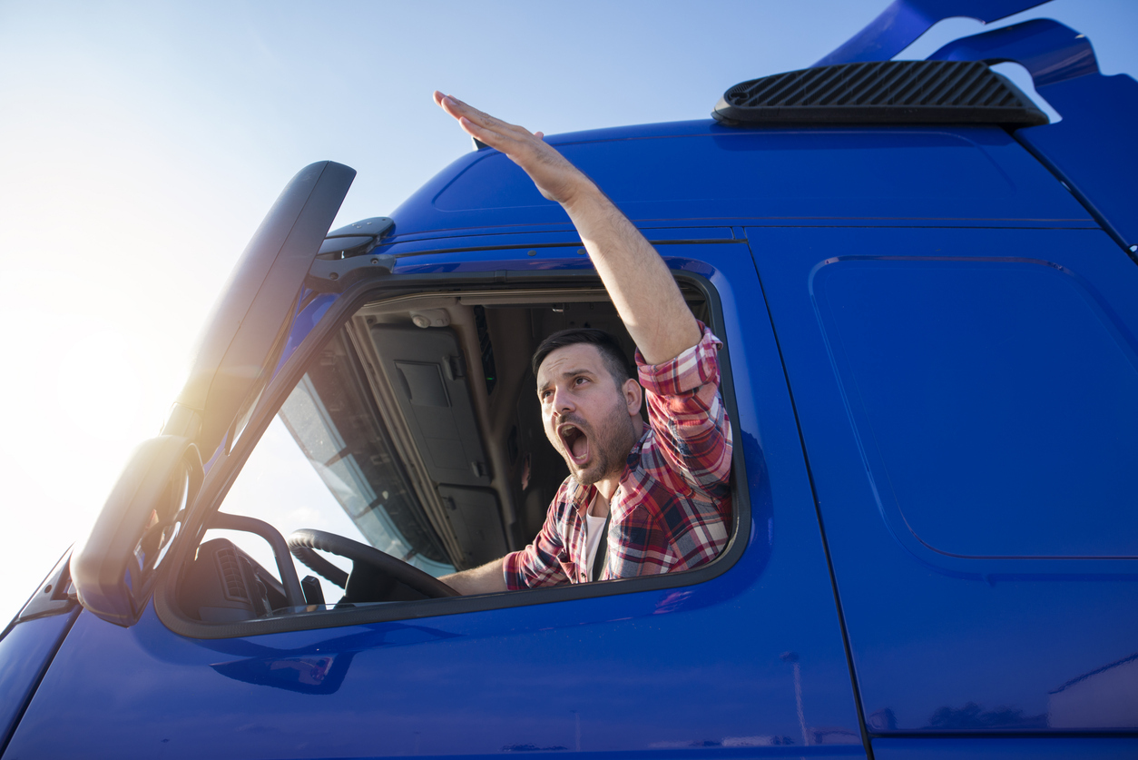 Truck driver leaning out of a blue semi-truck cab with one arm raised while on the road.