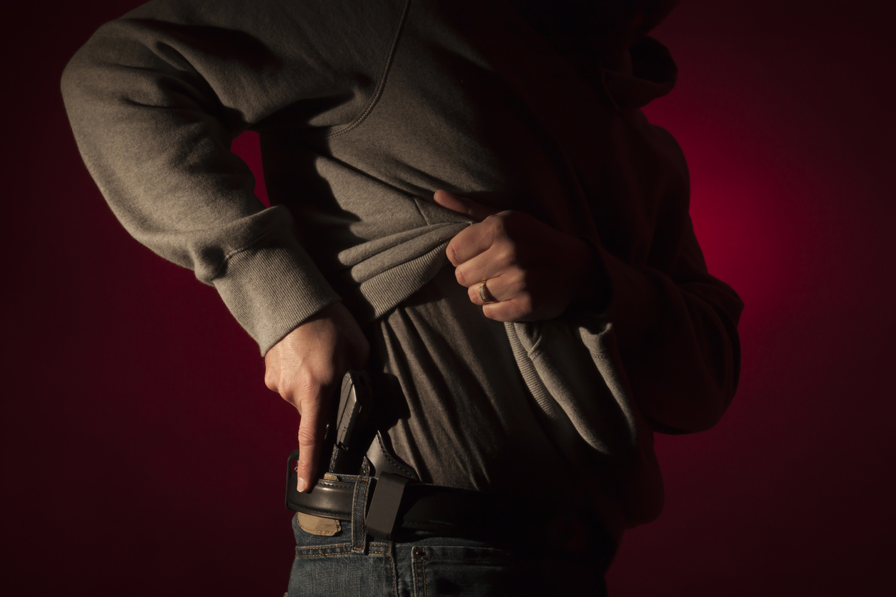 Person wearing a hoodie and jeans while carrying a concealed handgun in a waistband holster against a dark red background.