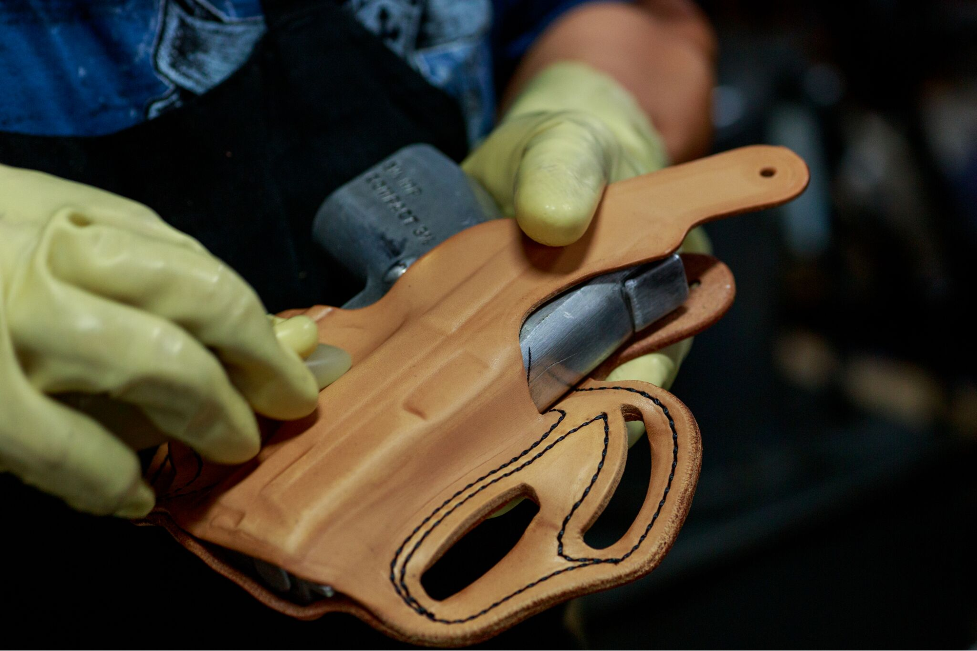 Close-up of a worker shaping a leather handgun holster around a metal mold while wearing protective gloves.